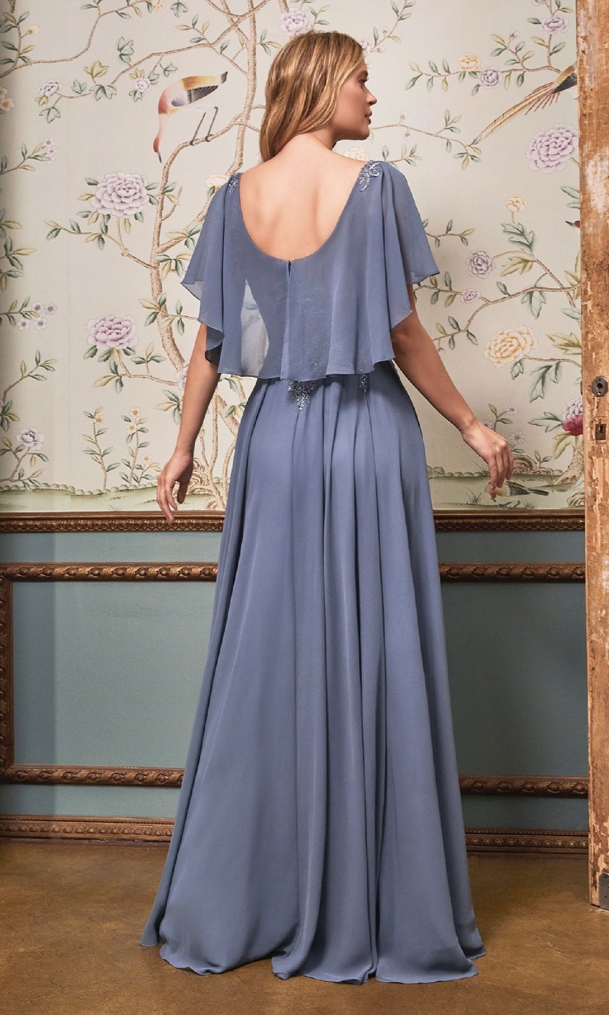 A woman in the Ladivine HT101 Cape-Sleeve Long A-Line Prom Dress, light blue chiffon with sheer short sleeves, stands facing a floral wallpapered wall.